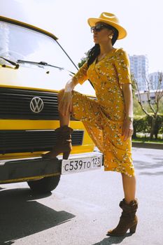 Stylish woman in floral dress and boots posing beside a yellow van on a sunny day.