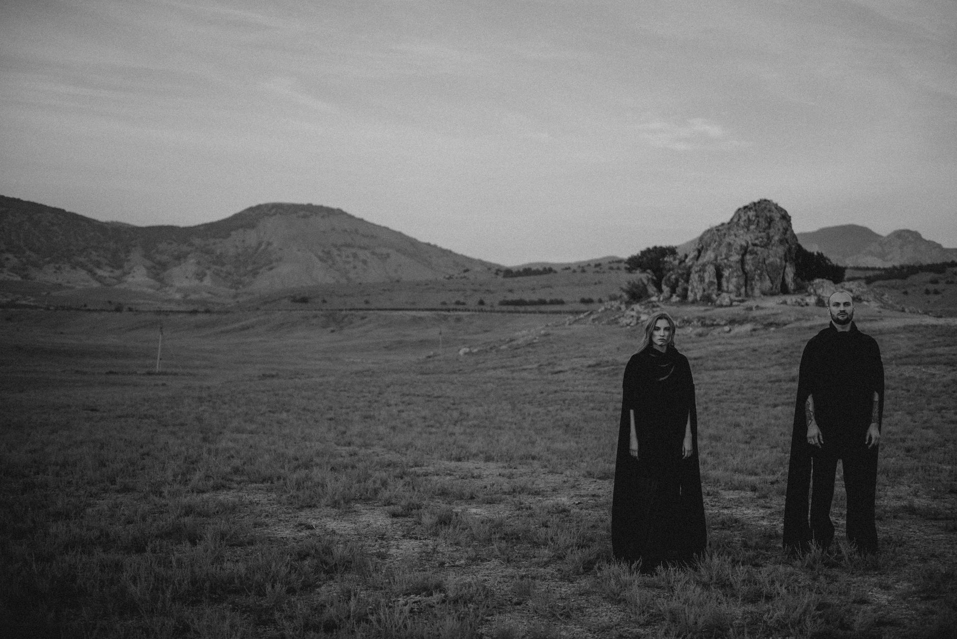 A solemn couple dressed in dark clothes stand in a vast, mountainous landscape.