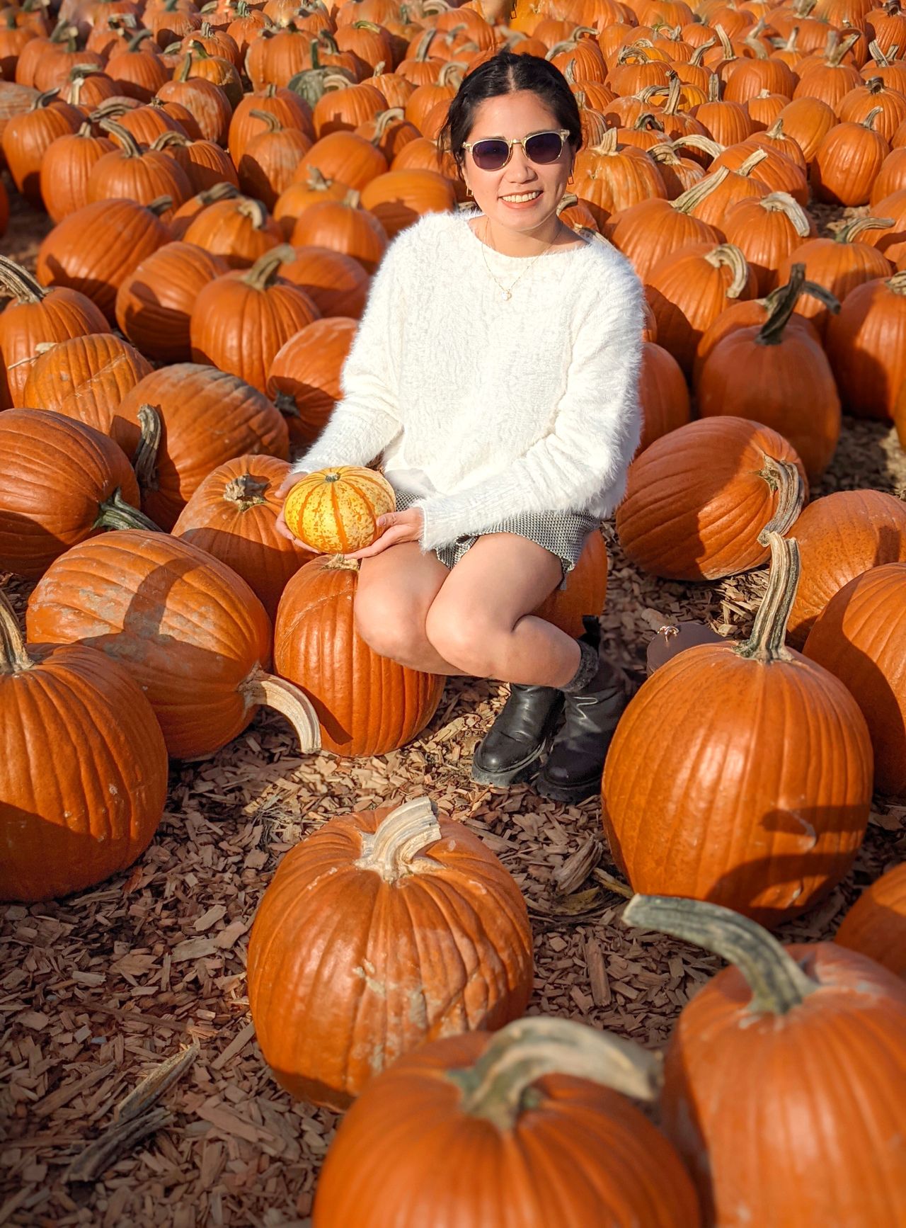 Woman in a soft white fuzzy sweater and black ankle boots crouching among pumpkins holding a small gourd