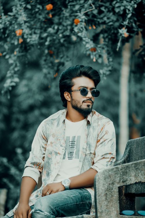 Free Fashionable man with sunglasses sitting on a park bench surrounded by greenery. Stock Photo