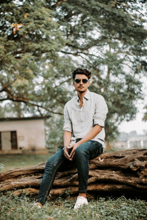 Free Young man in sunglasses sits confidently on a large tree log, embodying modern style. Stock Photo
