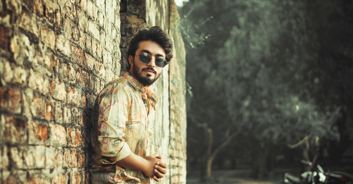 A man in a plain fitted top and sunglasses leaning against a brick wall, understated and clean