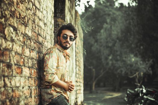 Man with sunglasses leaning against a brick wall, outdoors amidst trees.