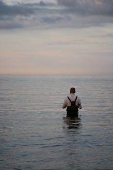 A lone fisherman stands in calm water at dawn, showcasing peaceful solitude and hobby.