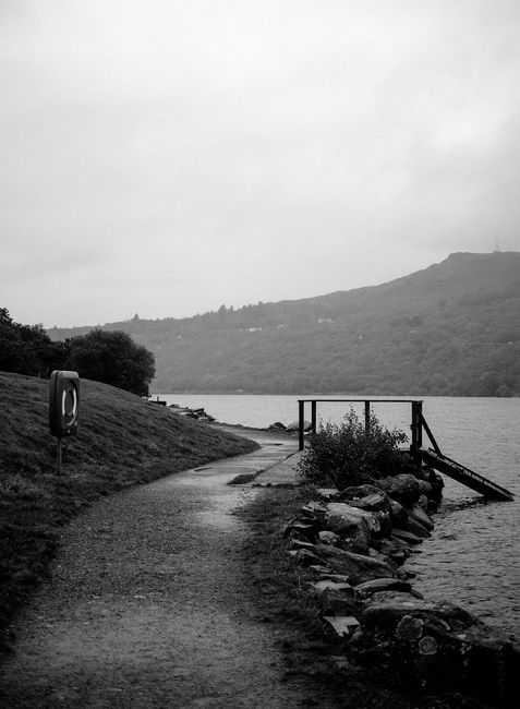Black and white image of a serene path along Llanberis Lake, Wales, with mountains in the background.