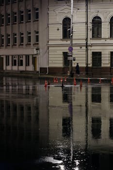City street during rain with building reflections at night, highlighting urban solitude.