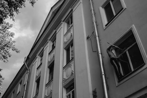 Grayscale Photo of a Man Cleaning the Glass Window of an Apartment Building