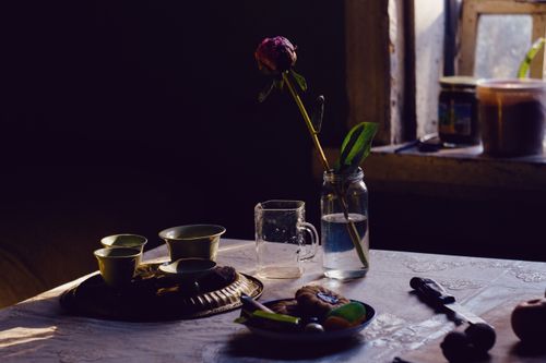 Free A serene indoor setting with a rose in a jar, cups, and tableware on a dimly lit table. Stock Photo