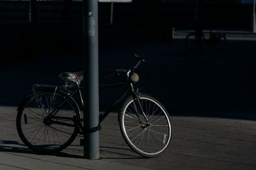 Free A bicycle is parked against a pole in a shadowy urban setting during daylight. Stock Photo