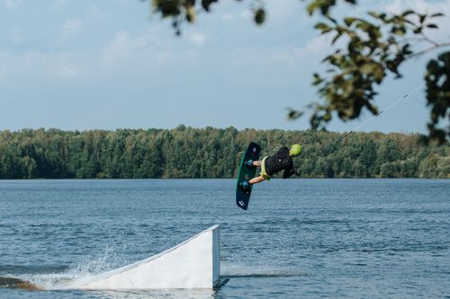 Free A wakeboarder performs a daring stunt mid-air over a ramp on a sunny lake, showcasing extreme sports prowess. Stock Photo