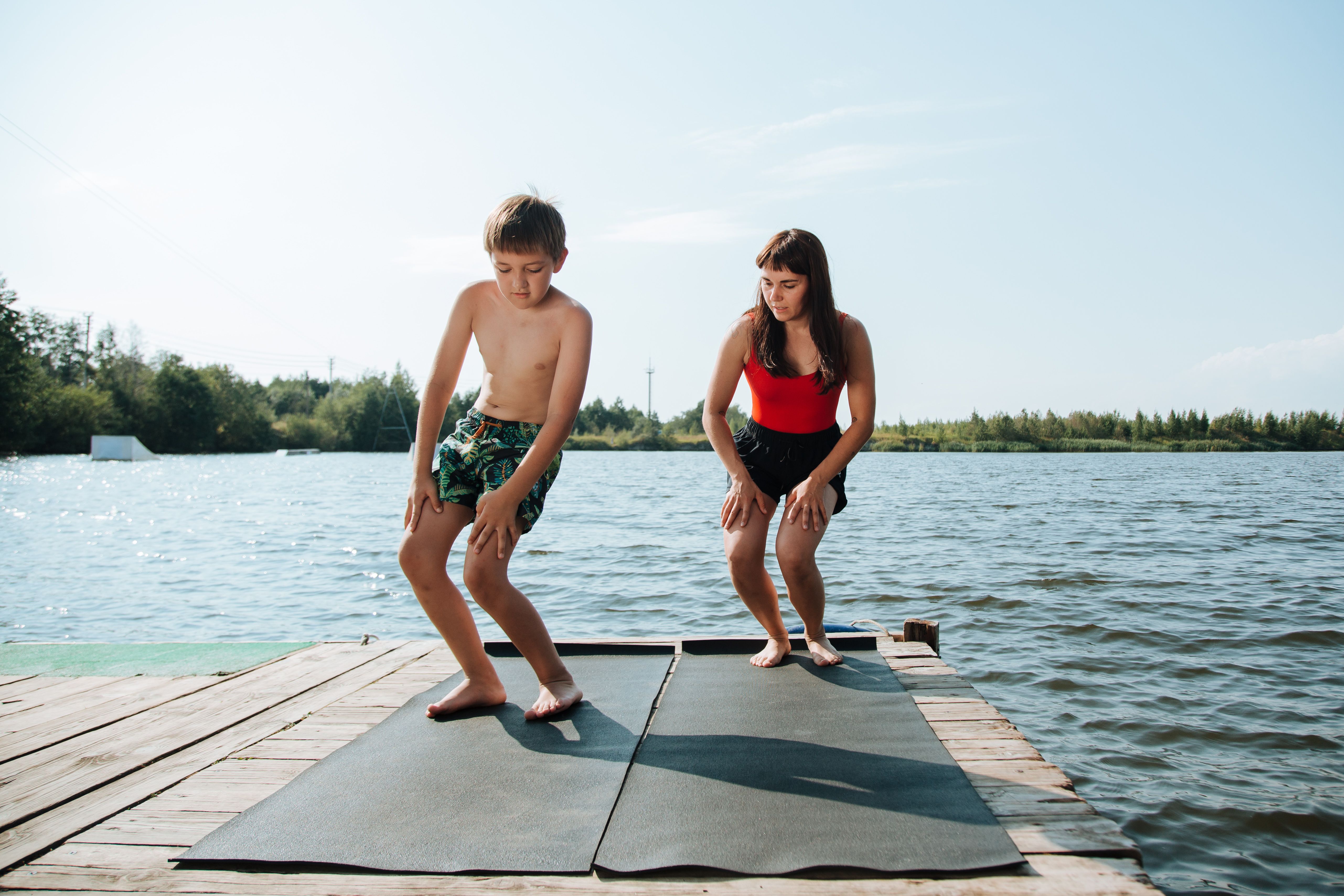 Mother and son stretch and warm up on a dock by the lake, enjoying a sunny day outdoor workout.