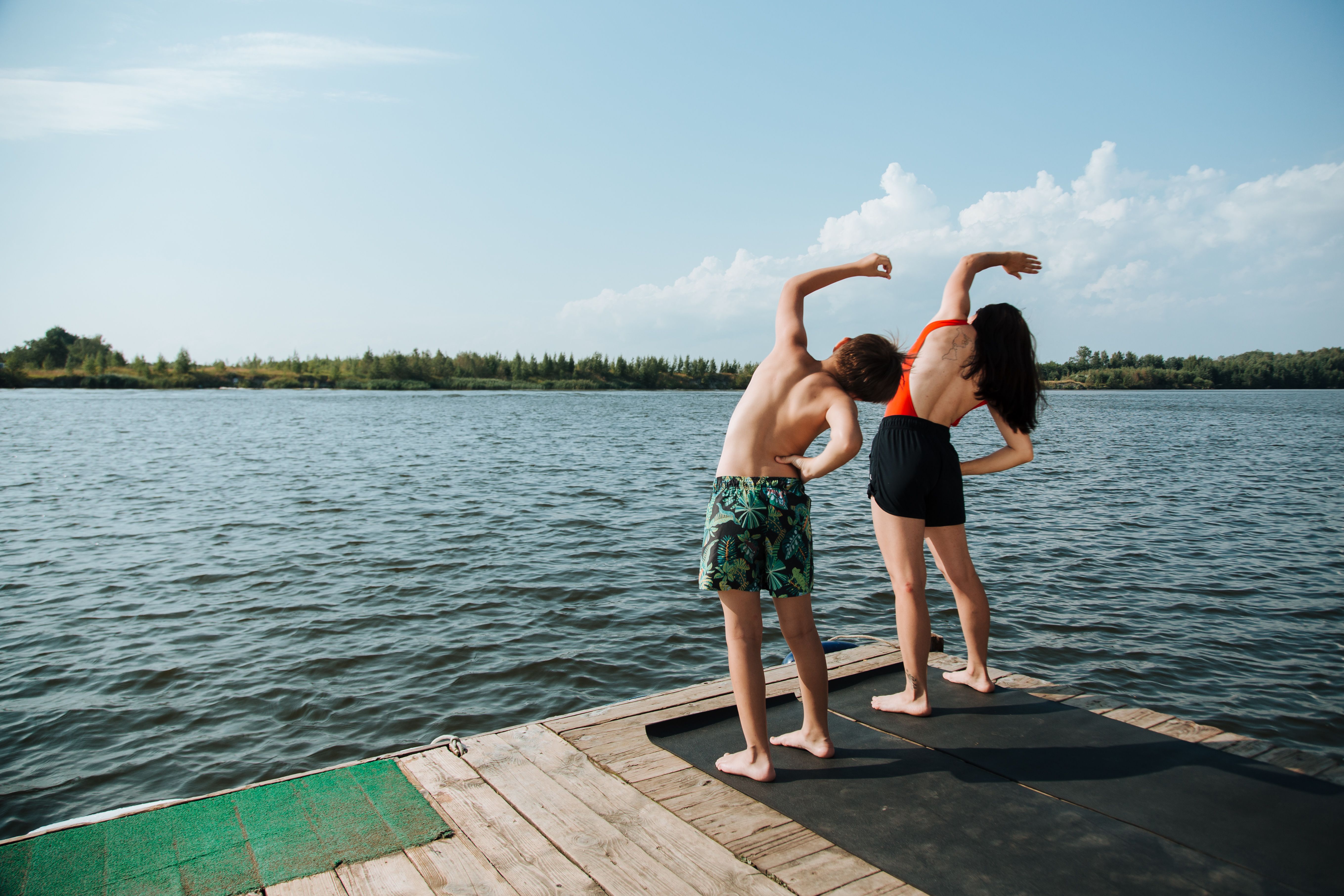 A mother and son stretch together on a lakeside dock under a clear summer sky.