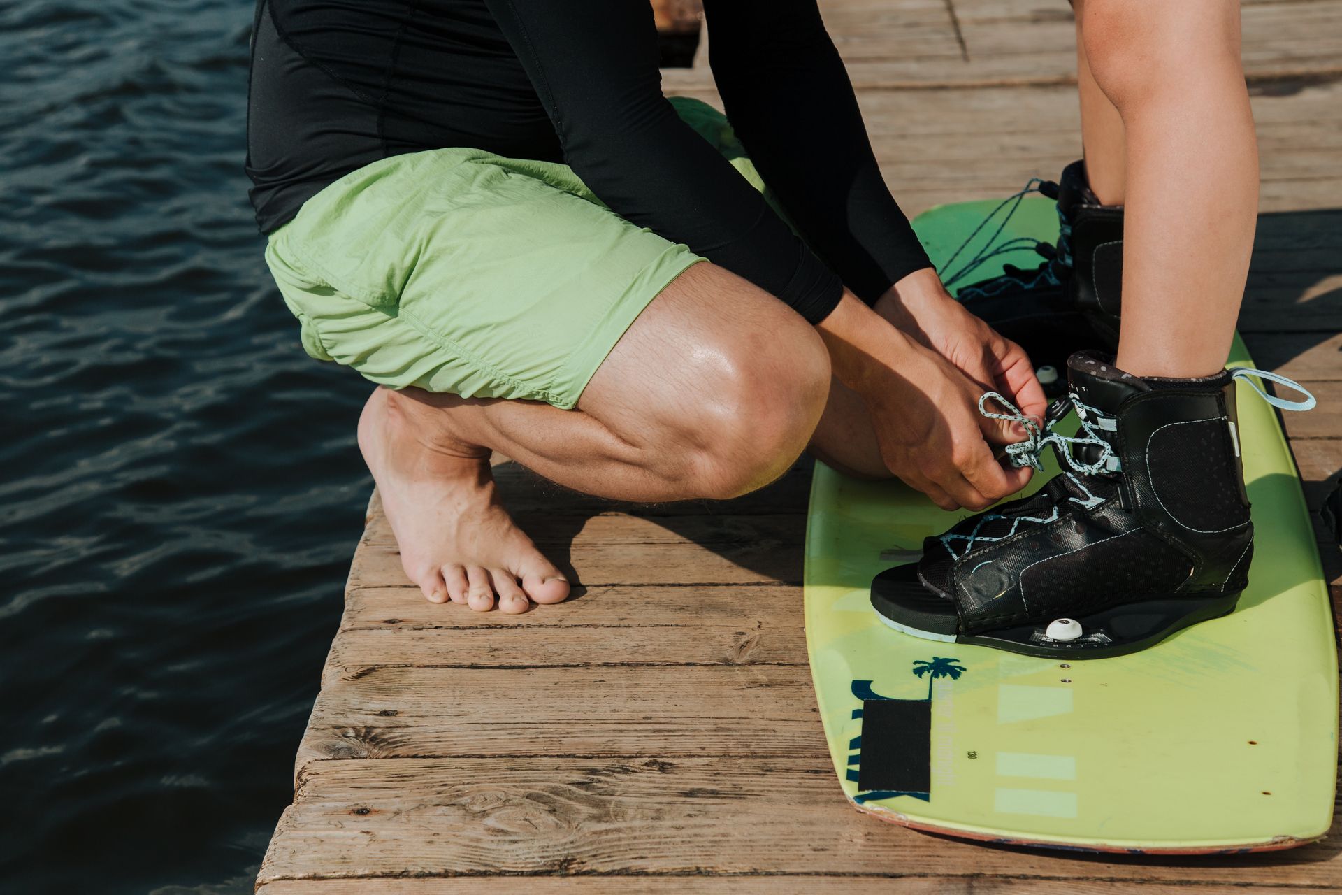A father helps his son prepare for wakeboarding on a lakeside jetty, ensuring safety and readiness.