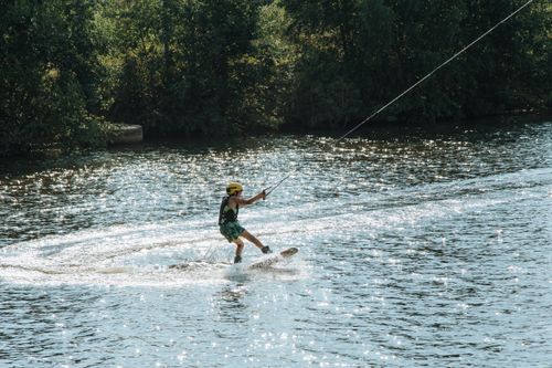 Free Young boy wakeboarding on a sunny day, wearing a helmet and vest, surrounded by nature. Stock Photo