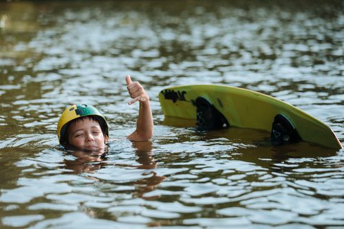 Free A child in a helmet gives a thumbs-up while wakeboarding in a calm lake, enjoying a summer day outdoors. Stock Photo