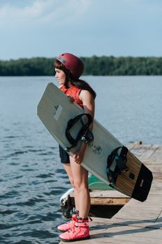 A woman in a helmet stands on a pier, holding a wakeboard, ready for an adventure on the lake.