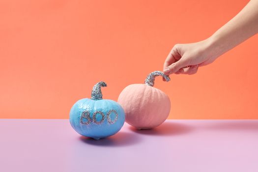 Hand holding pink pumpkin with glitter stem next to blue pumpkin with 'boo' text, orange background.