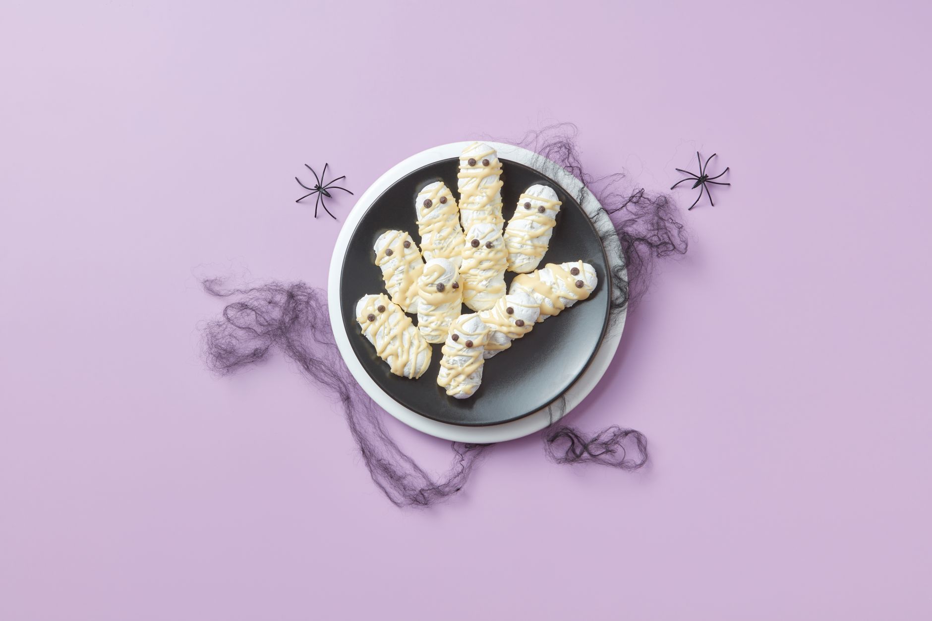 Creative Halloween themed cookies shaped like mummies on a purple background with decorative spiders.