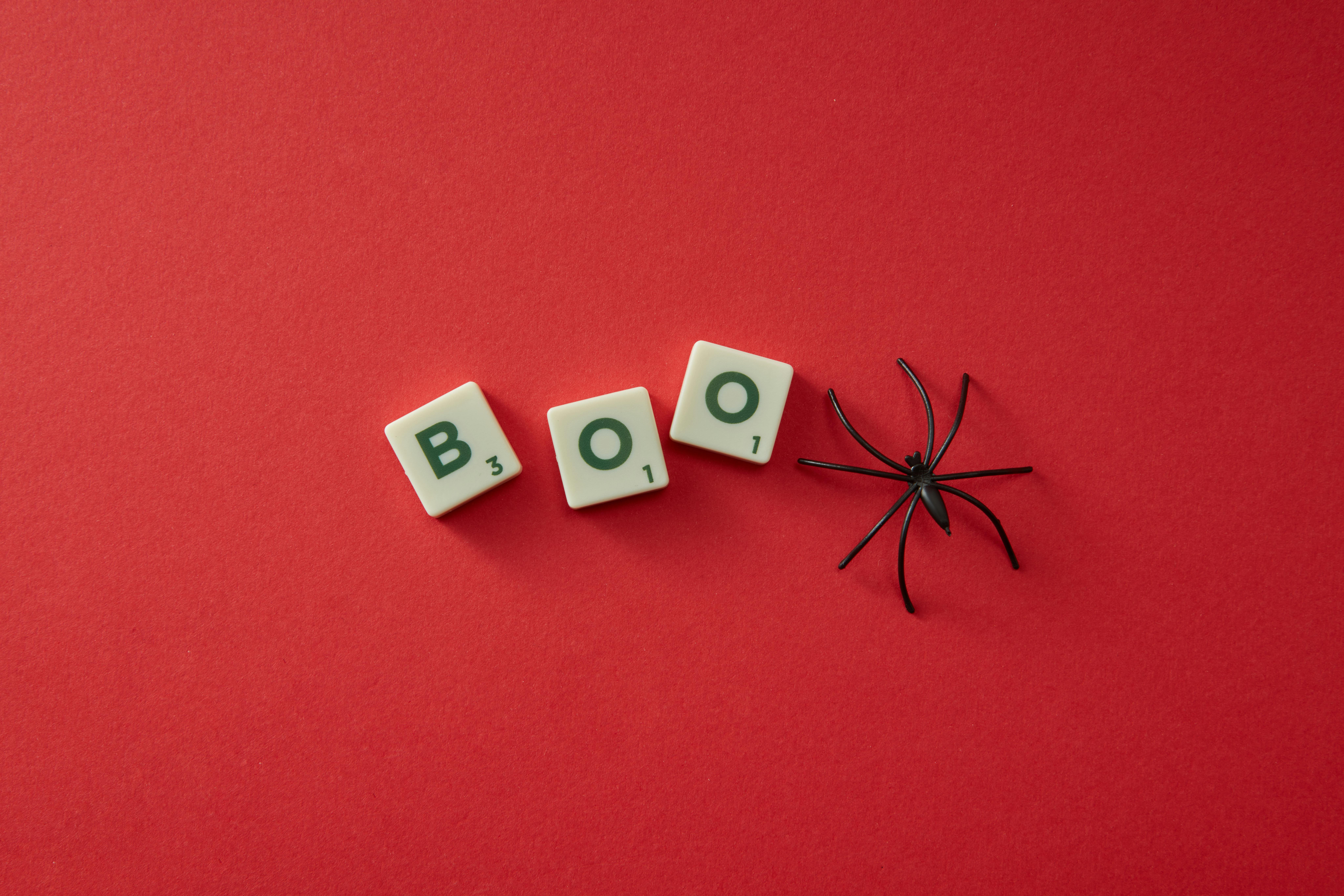 Scrabble tiles spelling 'BOO' with a black plastic spider on a red background, perfect for Halloween themes.