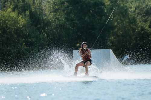 Free A woman wakeboarding skillfully on a sunny day with splashes of water and greenery in the background. Stock Photo