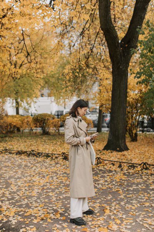 Free Asian woman in beige trench coat using smartphone in a fall park surrounded by leaves and trees. Stock Photo