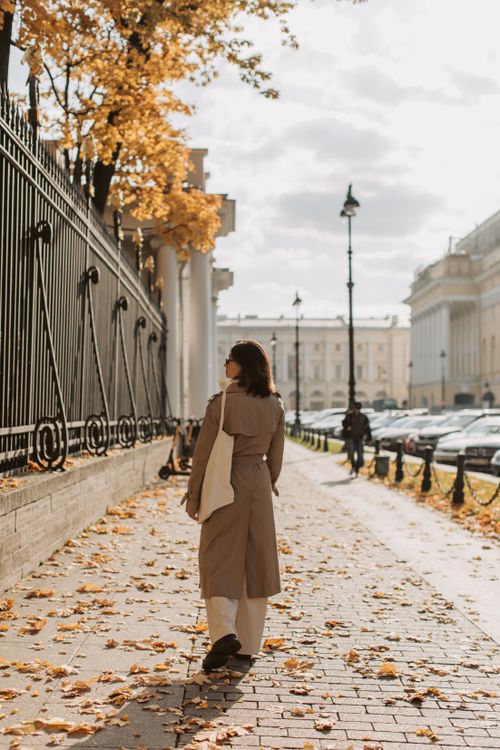 Free A woman in a beige coat walks along a leaf-strewn street in autumn, surrounded by buildings and a wrought iron fence. Stock Photo