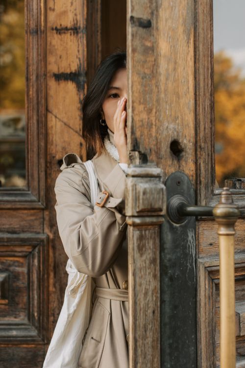 Free Asian woman in a trench coat peeks behind a rustic wooden door, evoking curiosity and mystery. Stock Photo