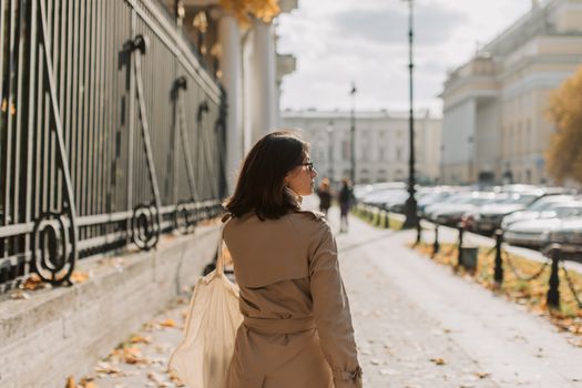 A woman in a trench coat walks along an urban sidewalk next to a metal fence on a sunny day.