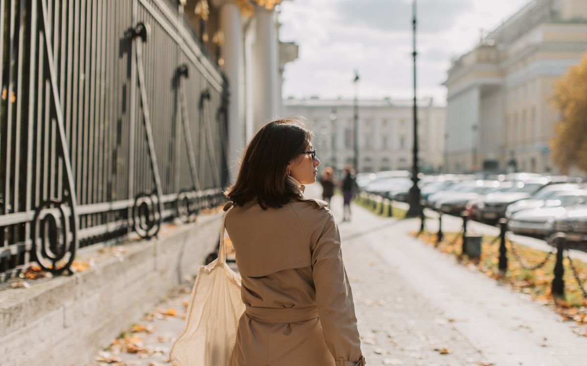 A woman in a trench coat walks along an urban sidewalk next to a metal fence on a sunny day.