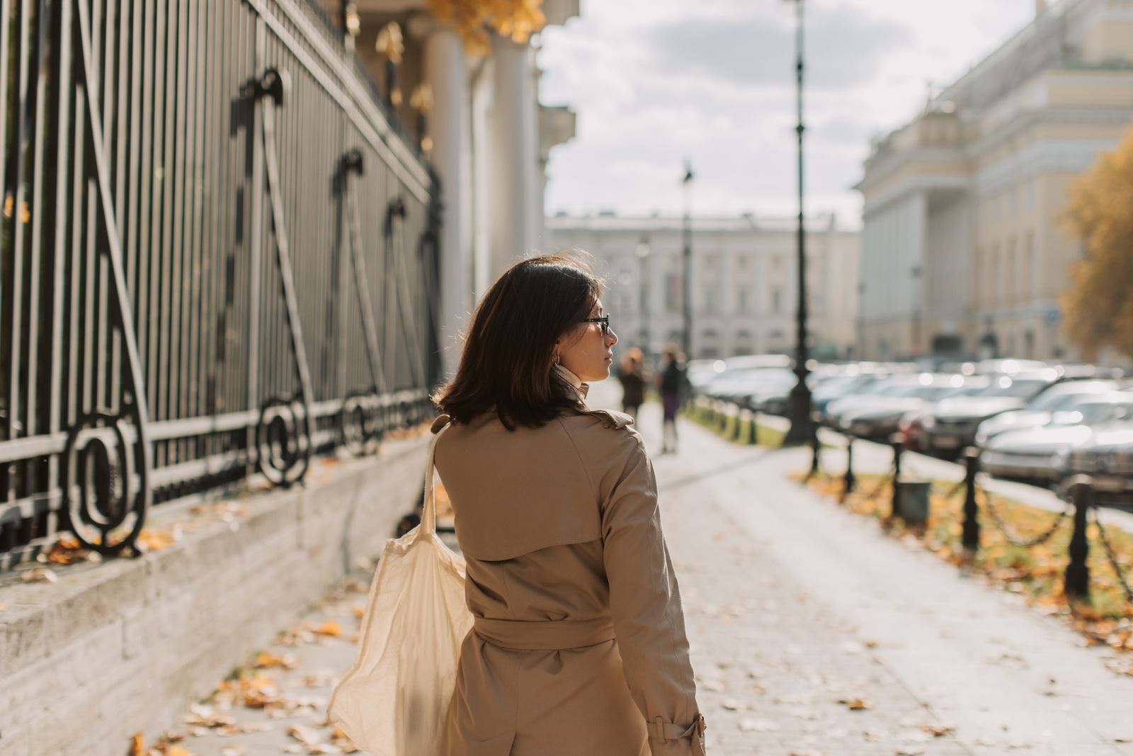 Woman in a beige trench coat walking along a sunlit autumn sidewalk beside an iron fence