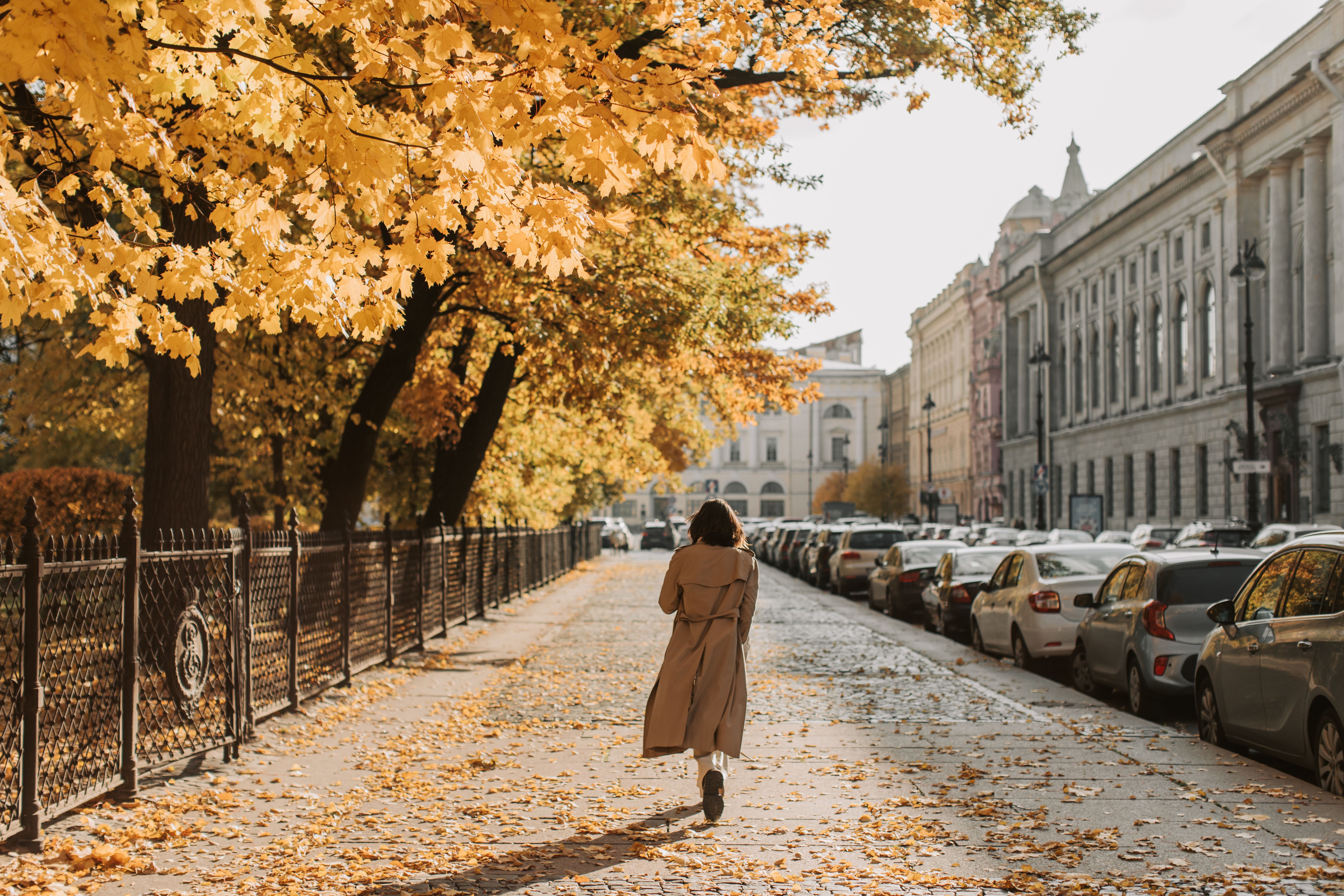 Woman in a trench coat walking a leaf covered city sidewalk past stone row houses in fall