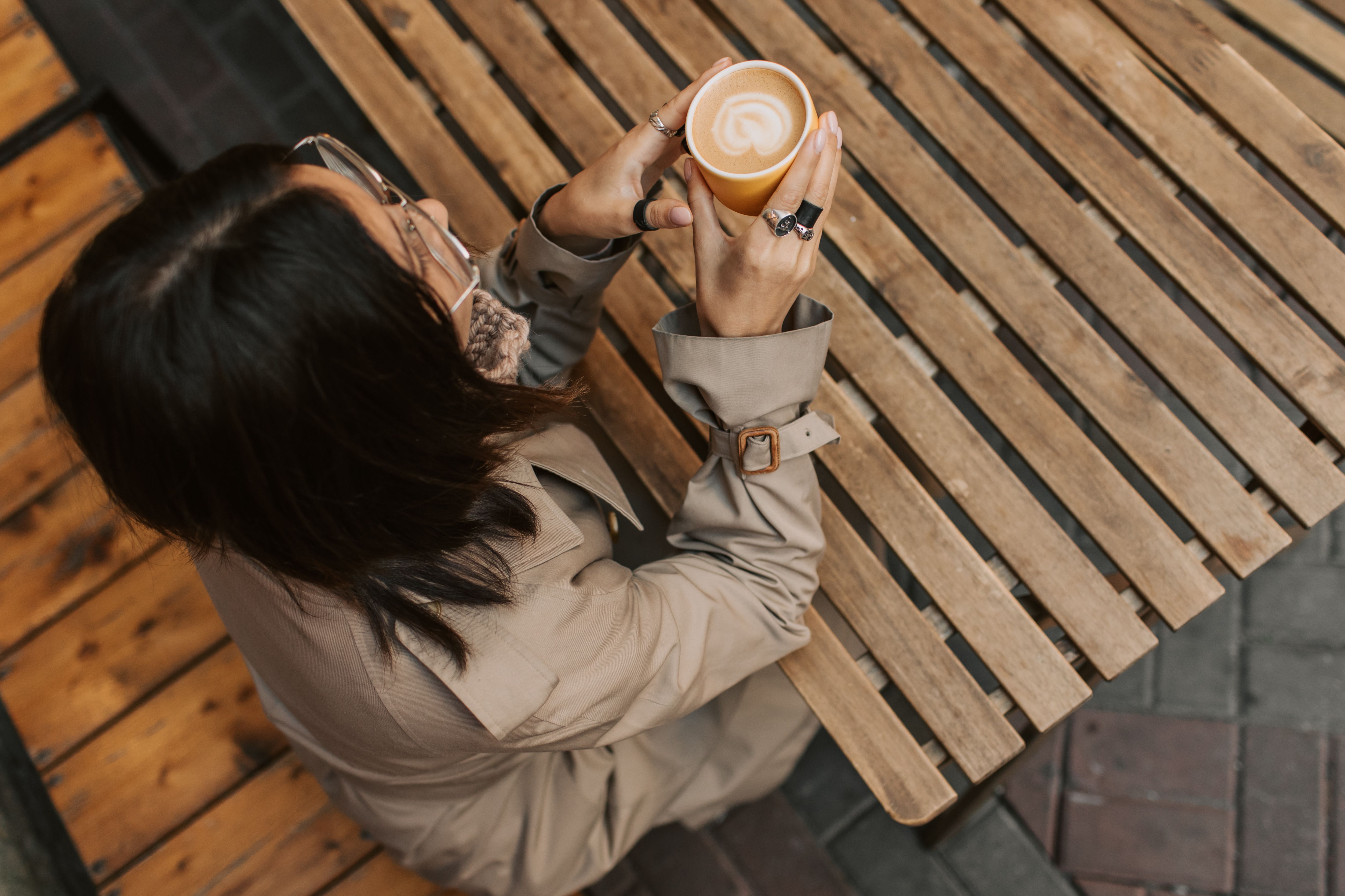A woman sitting at an outdoor cafe holding a latte in a beige coat.