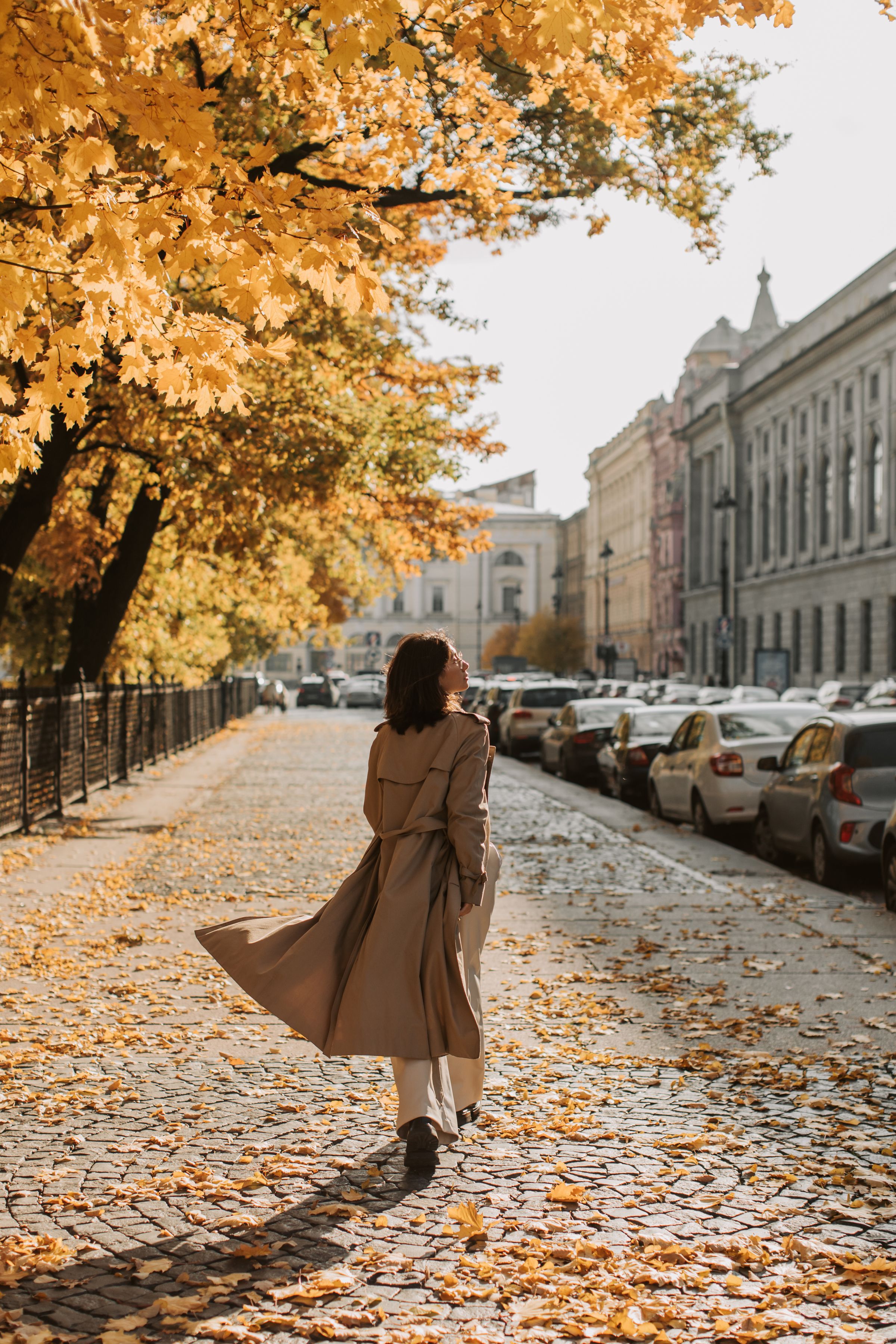 Woman in a trench coat walking on a tree-lined sidewalk in cool fall weather