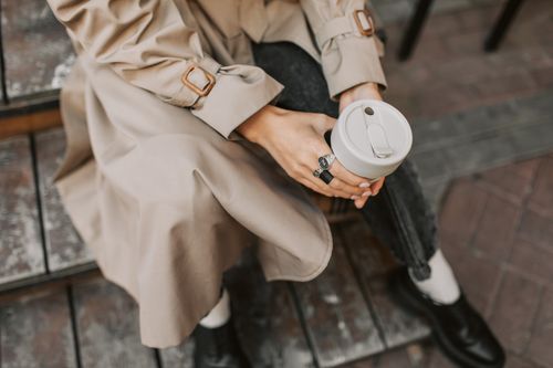 Free Close-up of a person holding a reusable cup while sitting, wearing a stylish trench coat. Stock Photo