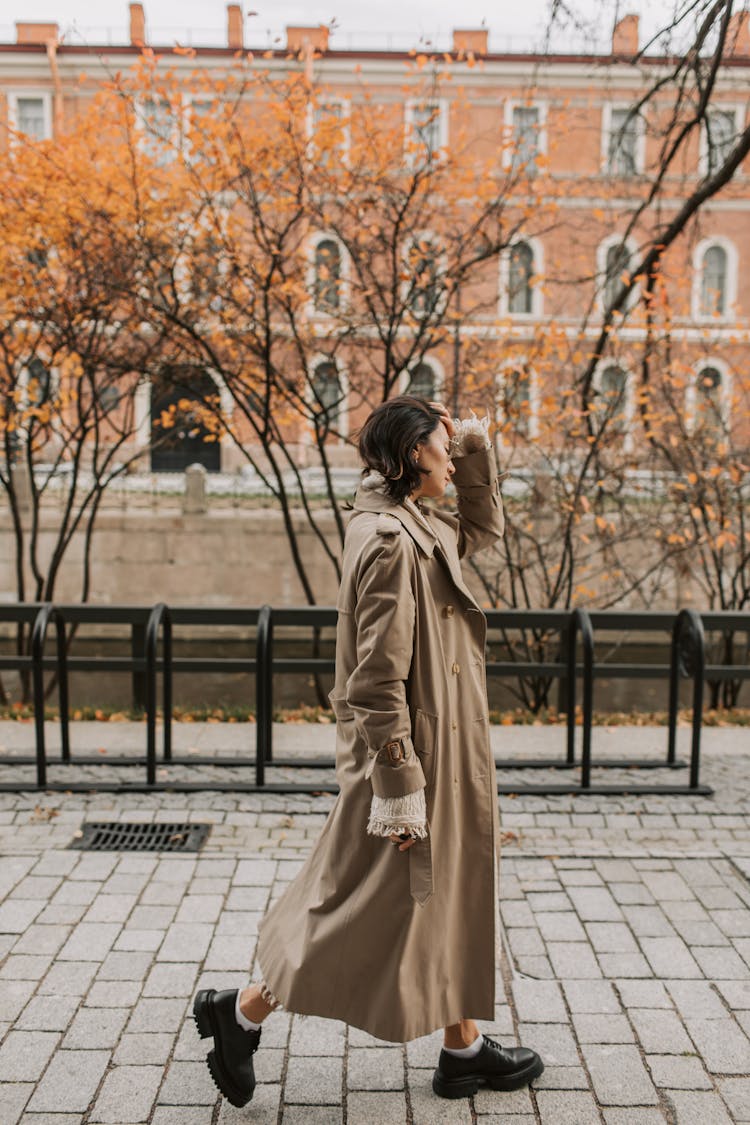 A Woman In Brown Trench Coat Walking On The Sidewalk