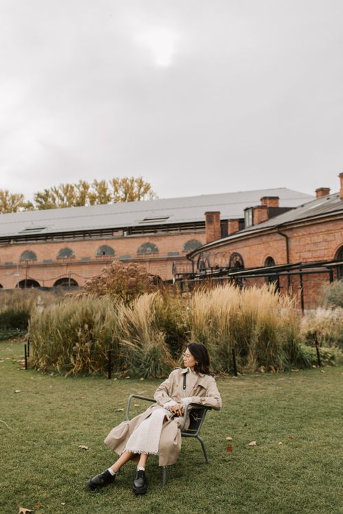 Free A woman in a trench coat sits outdoors in an autumn garden against a historical brick building. Stock Photo