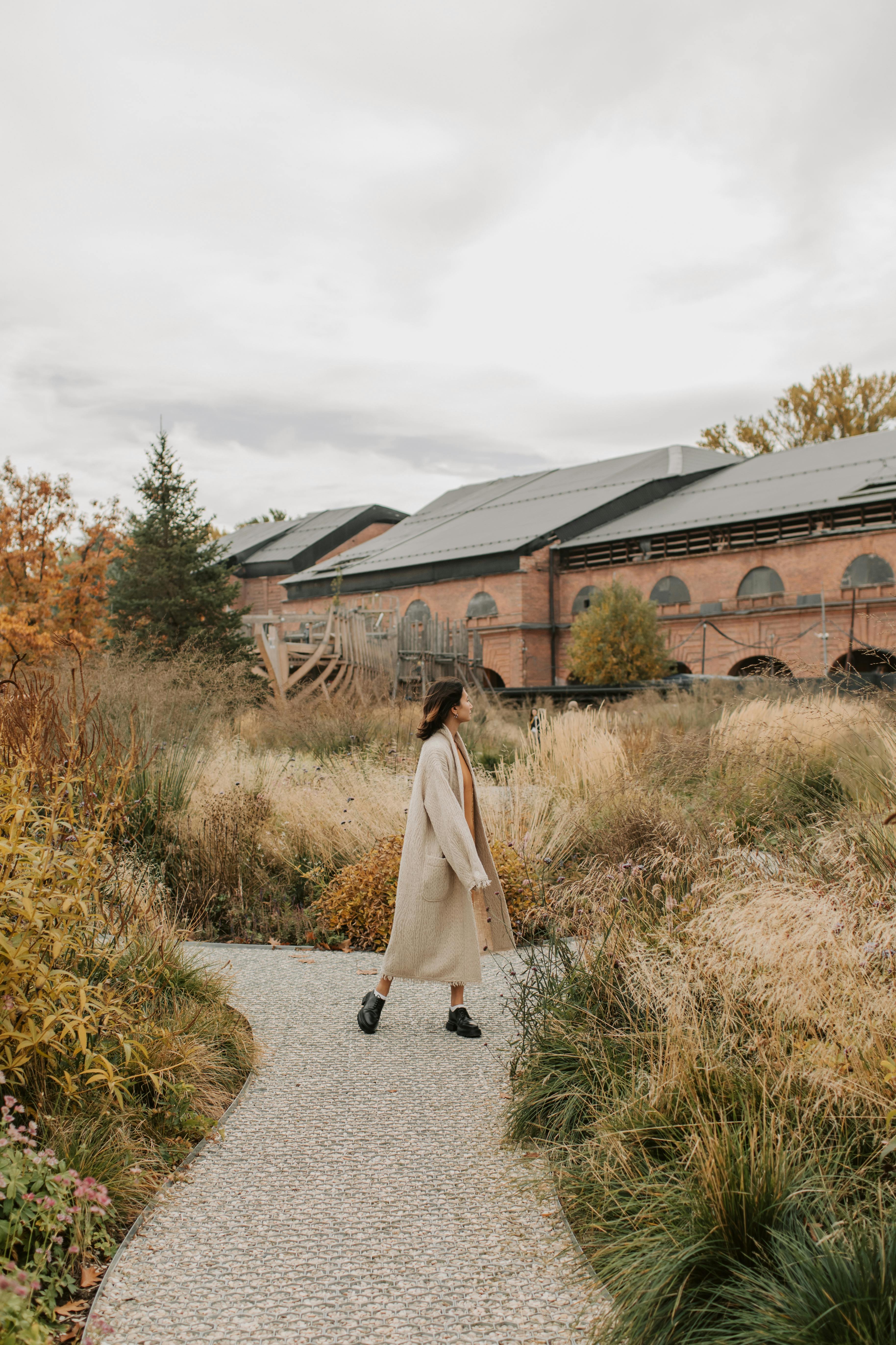 Woman in a trench coat walking on a path by a brick building in an autumn landscape.