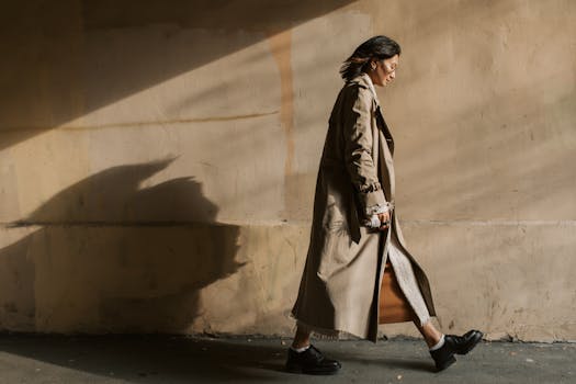 Stylish woman walking in sunlight casting shadows, wearing a trench coat against a textured wall.