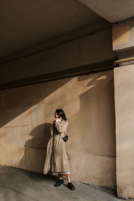 A fashionable woman posing in a beige trench coat, casting a shadow on a city street wall.
