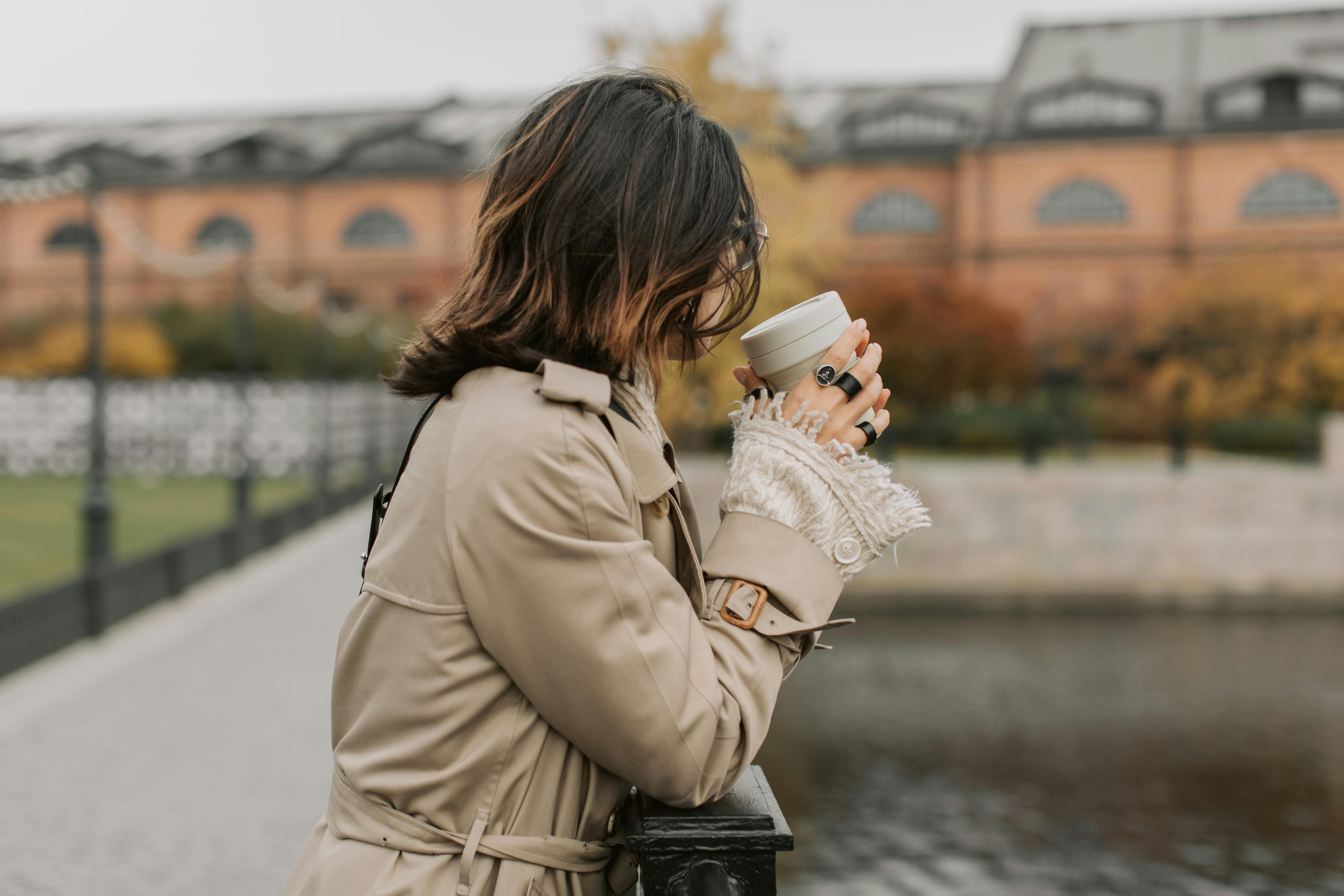A woman wearing a beige trench coat holds a cup by a peaceful river in an autumn setting.