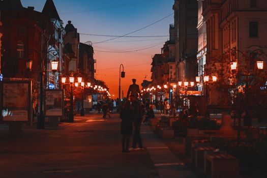 A bustling street scene at dusk, featuring warm illuminated lamps and silhouettes against a colorful sunset sky.
