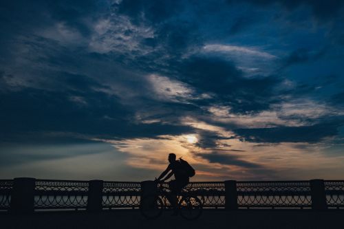 Free A person rides a bicycle silhouetted against a dramatic cloudy sunset sky, near a decorative railing. Stock Photo