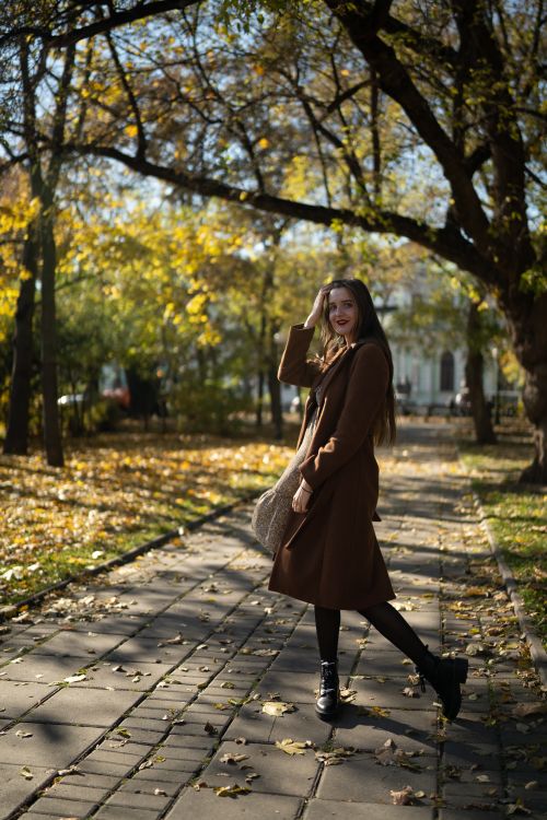 Free Woman in a brown coat smiles during an autumn walk along a leaf-covered pathway in Moscow. Stock Photo