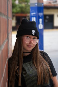 Portrait of a young woman with glasses and a beanie, leaning against a brick wall in daylight.