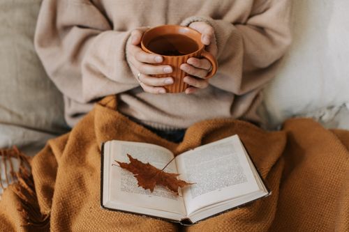 Free A woman enjoys a cozy autumn day, holding a tea mug over an open book with a maple leaf. Stock Photo