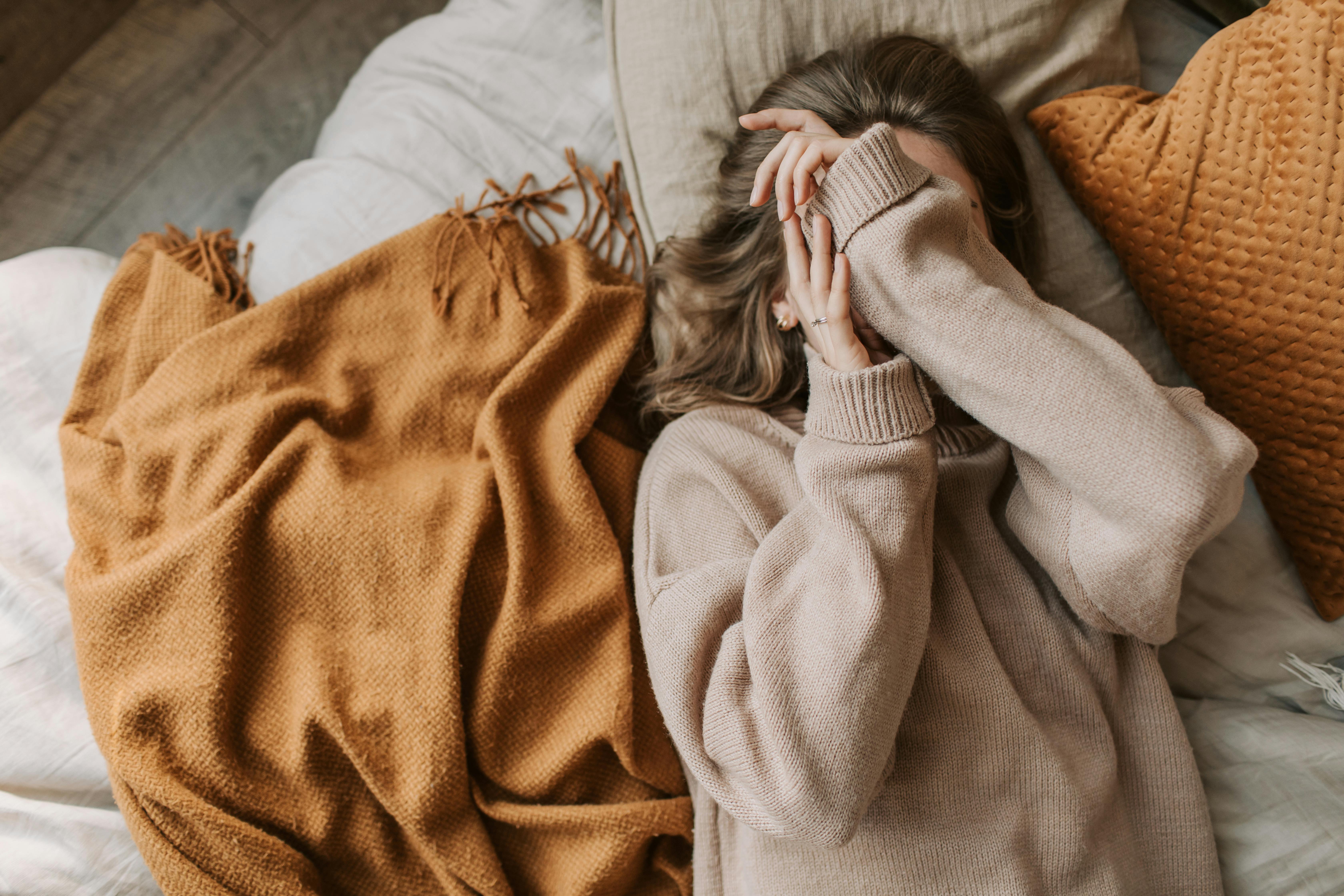Woman in knitted sweater relaxing on bed with cozy blankets, covering face for warmth.