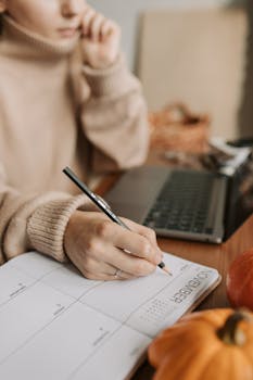 Close-up of a woman writing in a planner at home with a laptop and pumpkins.
