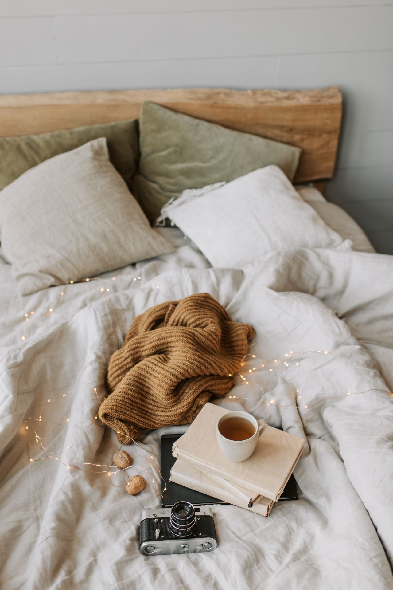 Bed with a mustard knit throw, fairy lights, stacked books, and a cup of tea