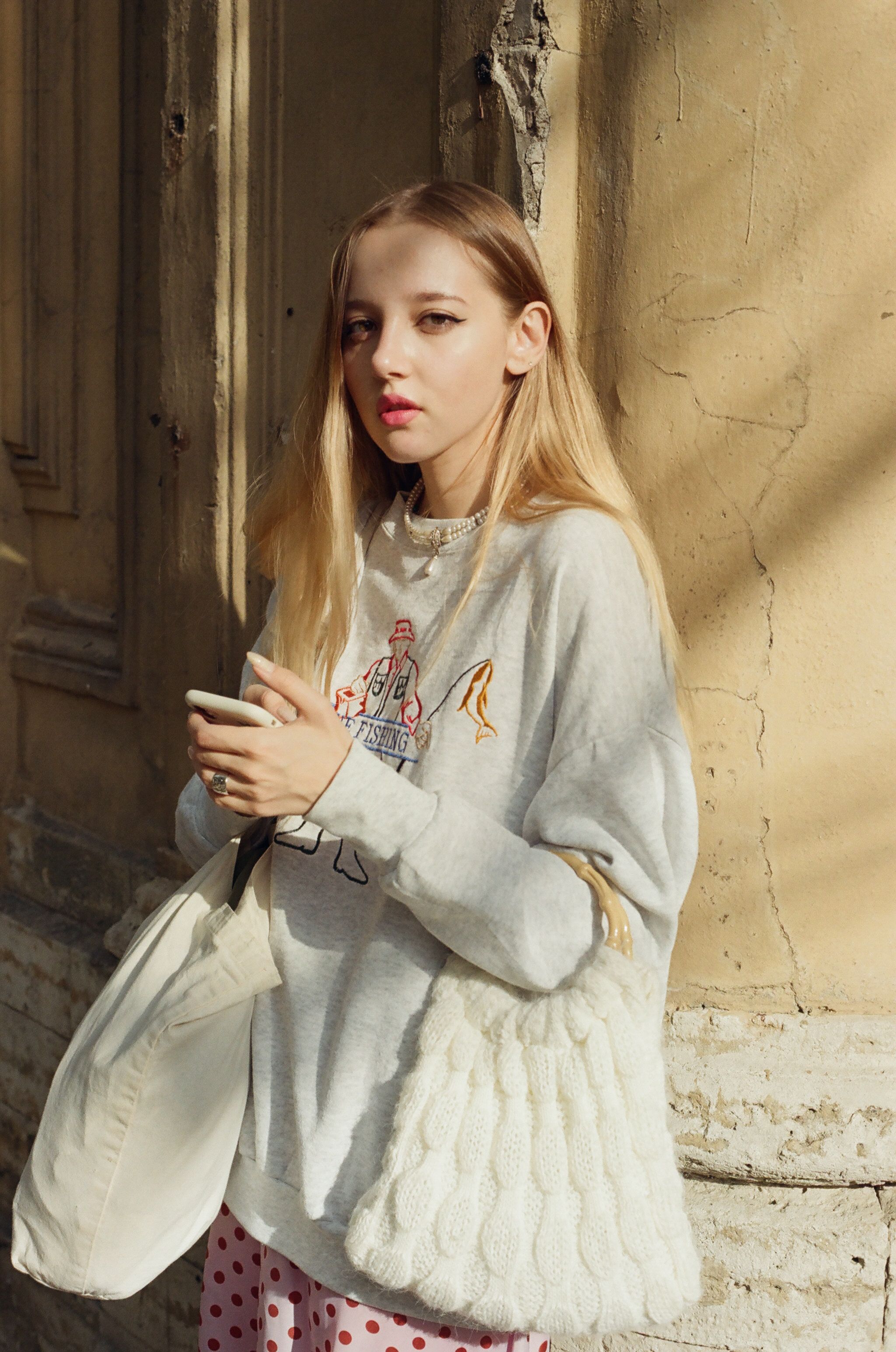 Young blond woman holding bags and posing against a textured wall outdoors.