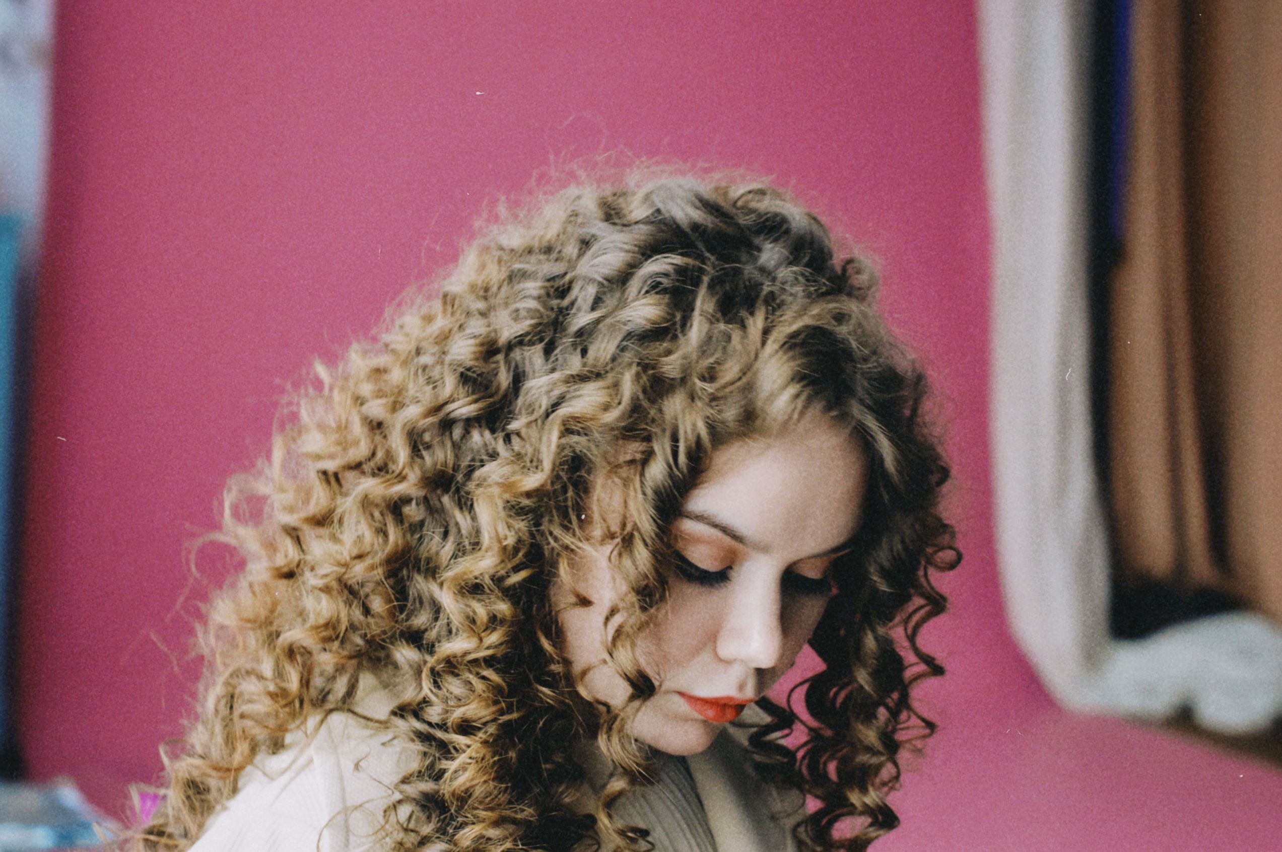 Close-up portrait of a woman with curly hair looking down against a pink backdrop.